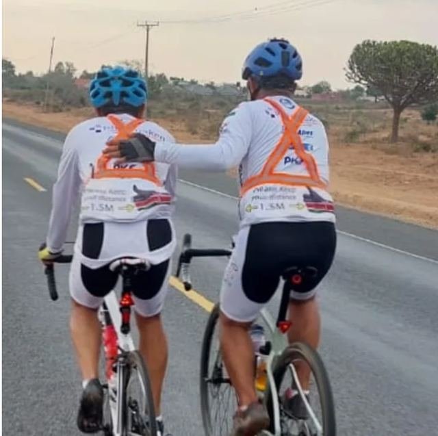Two riders on a Kenyan road, one resting a hand on the other's back